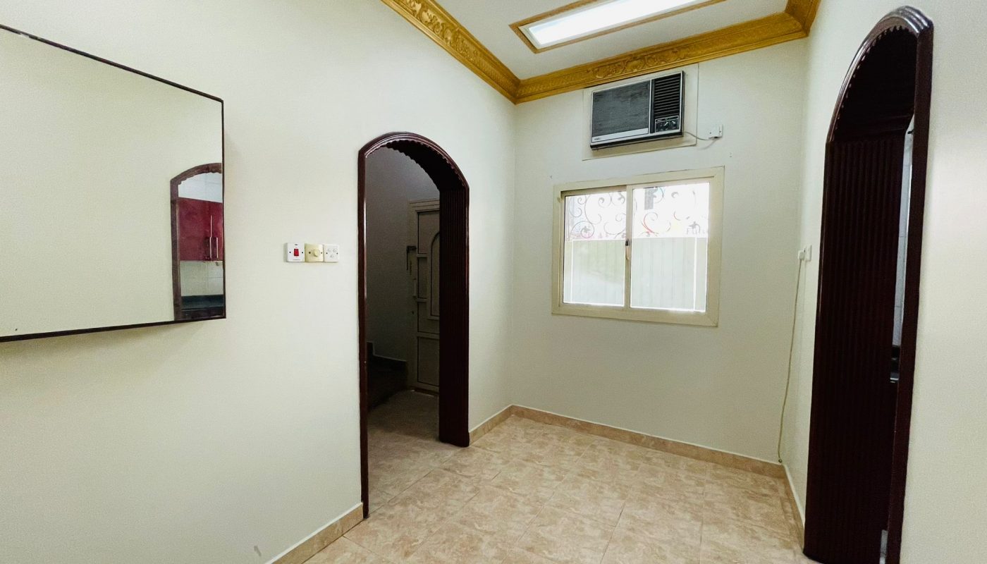 An empty room in a 2BR Villa located in Ras Rumman, featuring a tiled floor, white walls, an air conditioning unit, a window with decorative grills, and two doorways.