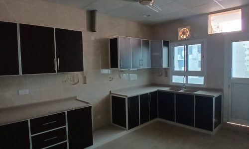 A dimly lit kitchen in a Hidd area apartment, featuring dark cabinets, a fridge, and a ceiling fan, with sunlight entering through a window.