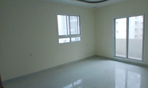 Empty, bright apartment room in a hidden area with white walls, shiny floor, and a corner window next to a ceiling fan.