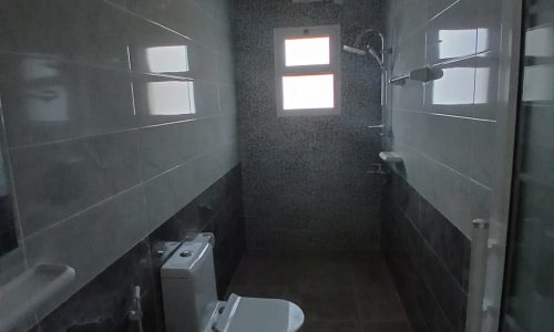 A dimly lit bathroom in a hidden area of the apartment for sale, with gray tiles, featuring a toilet on the left and a sink on the right, a mirror hanging above the sink, and