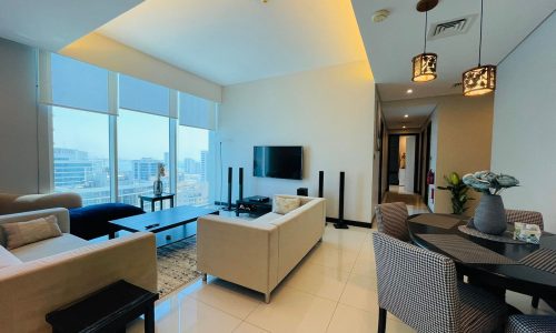 Luxury 2BR Apartment for rent in Juffair, featuring a modern living room with large windows overlooking the city, a sectional sofa, flat-screen TV, dining area, and warm lighting.