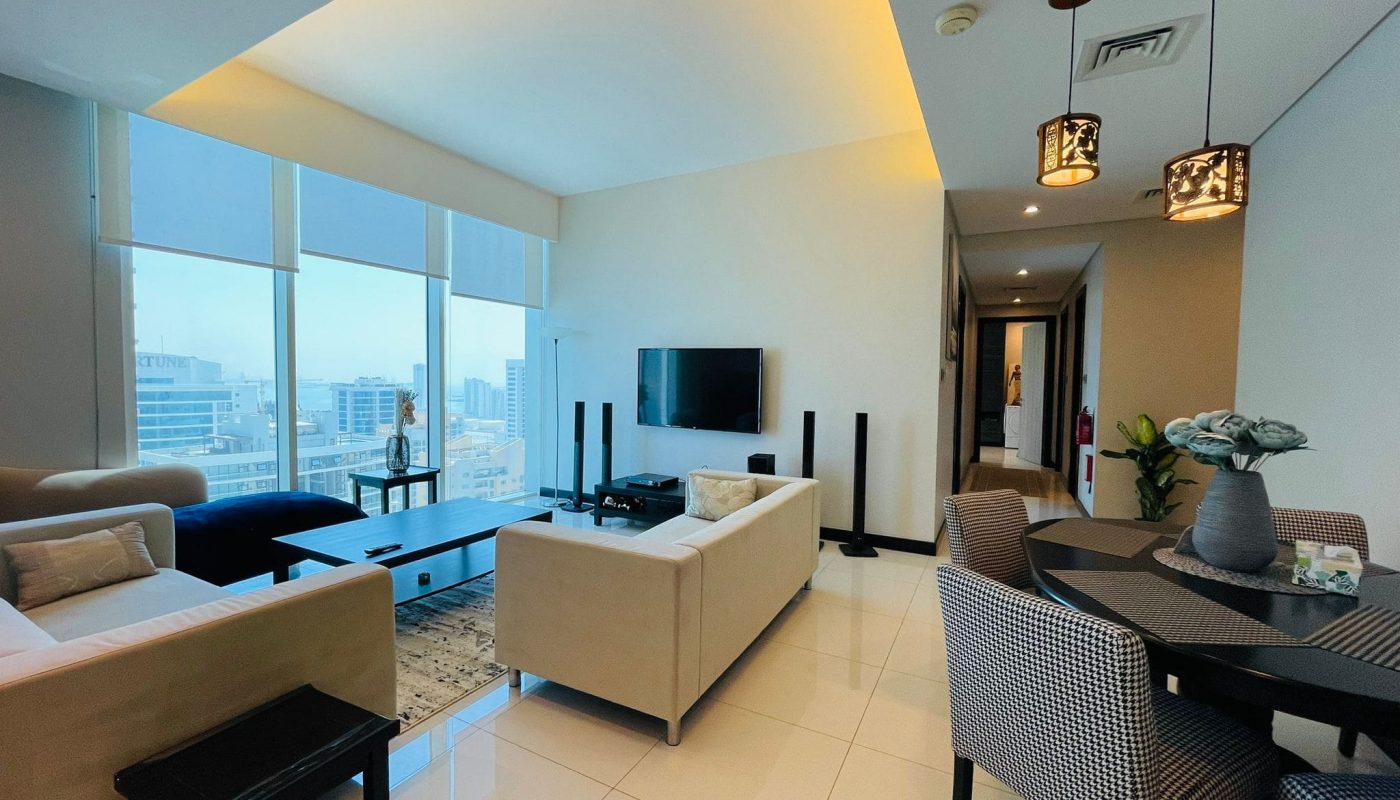 Luxury 2BR Apartment for rent in Juffair, featuring a modern living room with large windows overlooking the city, a sectional sofa, flat-screen TV, dining area, and warm lighting.