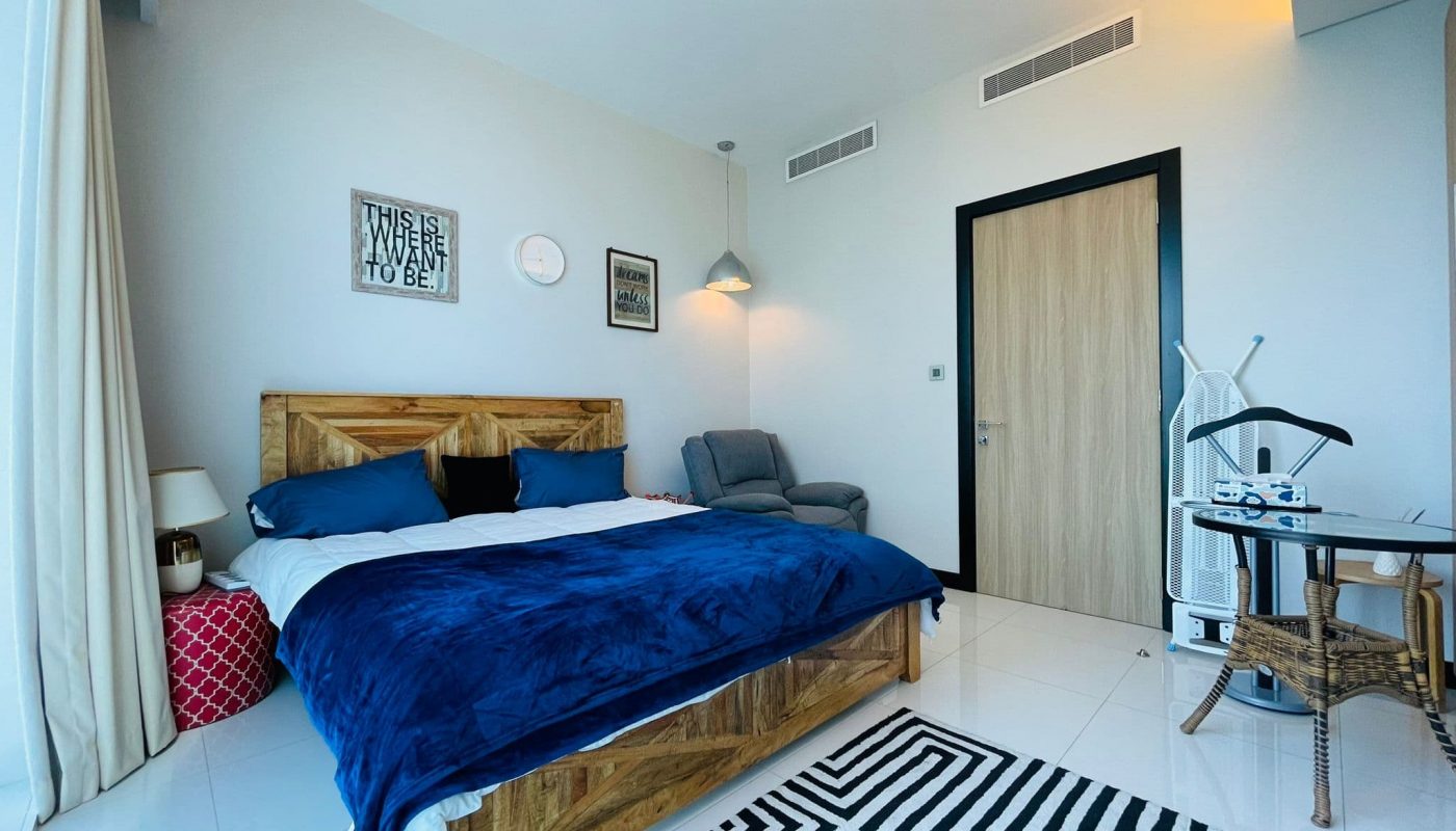 Modern 2BR bedroom in a Luxury Apartment, with a wooden bed, blue bedding, a gray armchair, and black and white decor accents in Juffair.