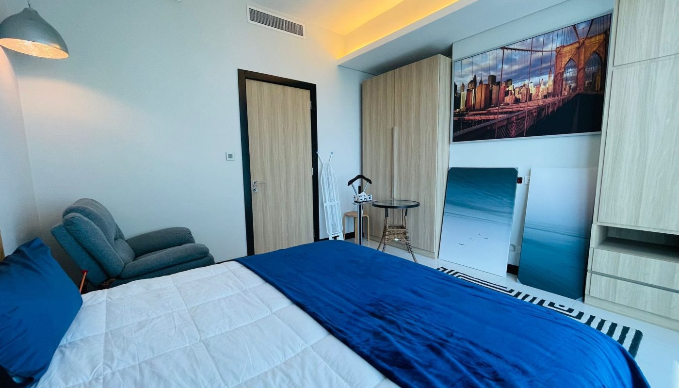Luxury 2BR apartment with a modern bedroom featuring a blue and white bedspread, work desk, exercise equipment, and a cityscape wall art.