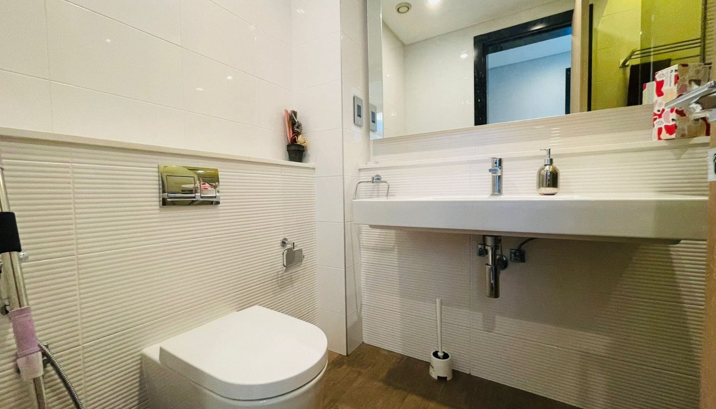 A modern bathroom in a luxury 2BR apartment with a white ceramic toilet and a floating sink with a large mirror.