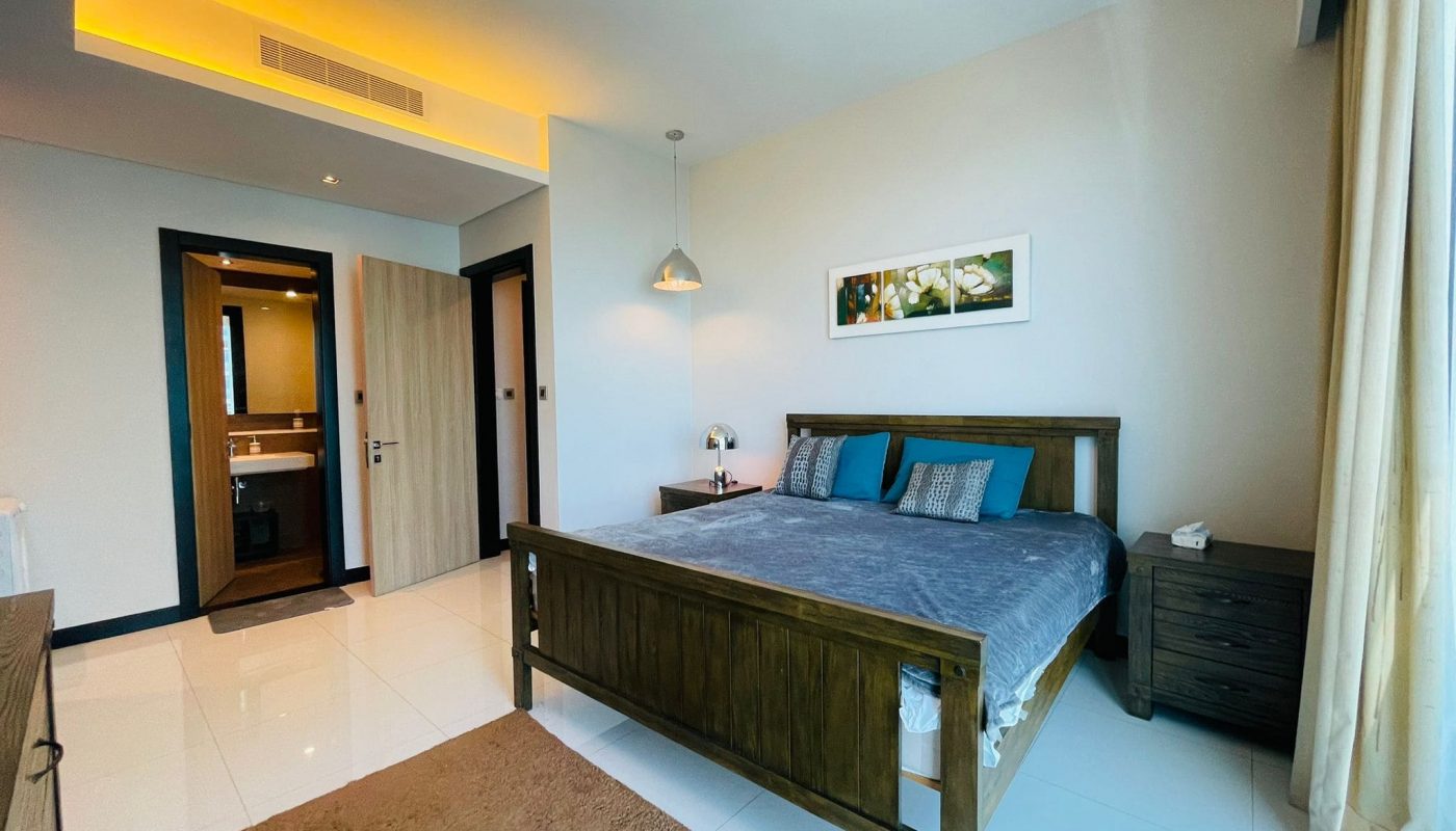 Luxury apartment bedroom in Juffair with a wooden bed, blue bedding, ambient lighting, and an attached bathroom.