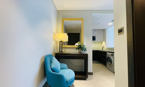 A modern hallway featuring a blue armchair, a console table with a lamp and flowers, a mirror, and an open door leading to a laundry area.