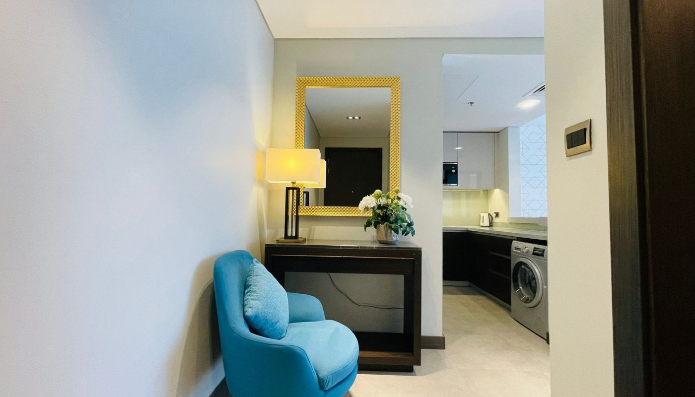 A modern hallway featuring a blue armchair, a console table with a lamp and flowers, a mirror, and an open door leading to a laundry area.