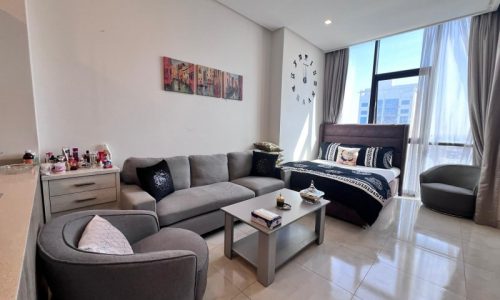 A modern, neatly arranged studio flat for sale in Juffair with a gray sectional sofa, matching armchair, and a bed with a dark headboard; a wall-mounted painting and decorative items are