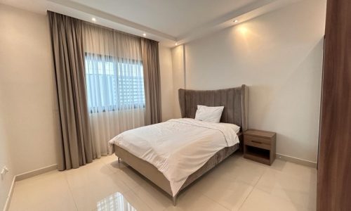 Modern minimalist bedroom in a luxury apartment with a neatly made bed, grey headboard, white bedding, and curtains at a window.