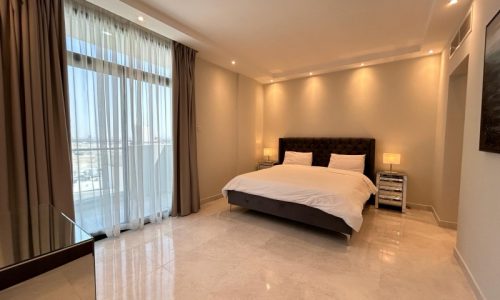 A modern bedroom in a luxury apartment, featuring a large bed with a tufted headboard, marble floors, and sheer curtains with city views.