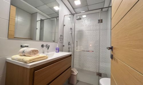 A luxury bathroom with a glass shower stall, wooden cabinetry, white sink, towel, and toiletries.