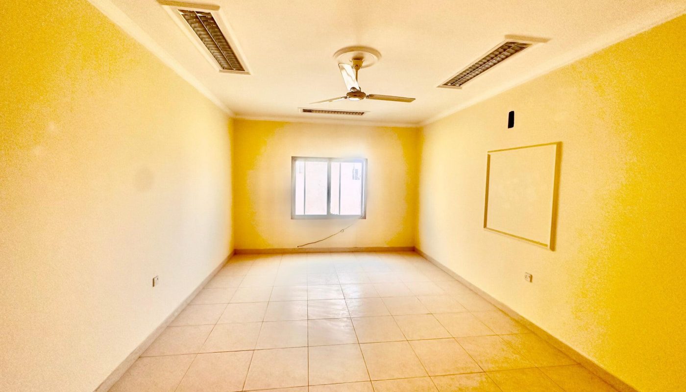An empty two-bedroom apartment in the Janbyah area boasts beige walls, a window, a ceiling fan with lights, rectangular light fixtures, and tile flooring. A small black object and a whiteboard are mounted on the walls. Ideal for rent with ample space to accommodate various living arrangements.