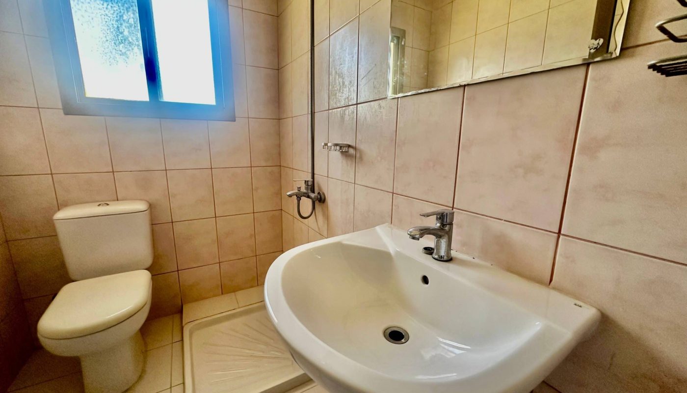 A small bathroom in a Janbyah, featuring beige tiled walls, a white sink, toilet, and shower area. The room includes a window with frosted glass allowing natural light to enter, perfect for those seeking two-bedroom apartments for rent.