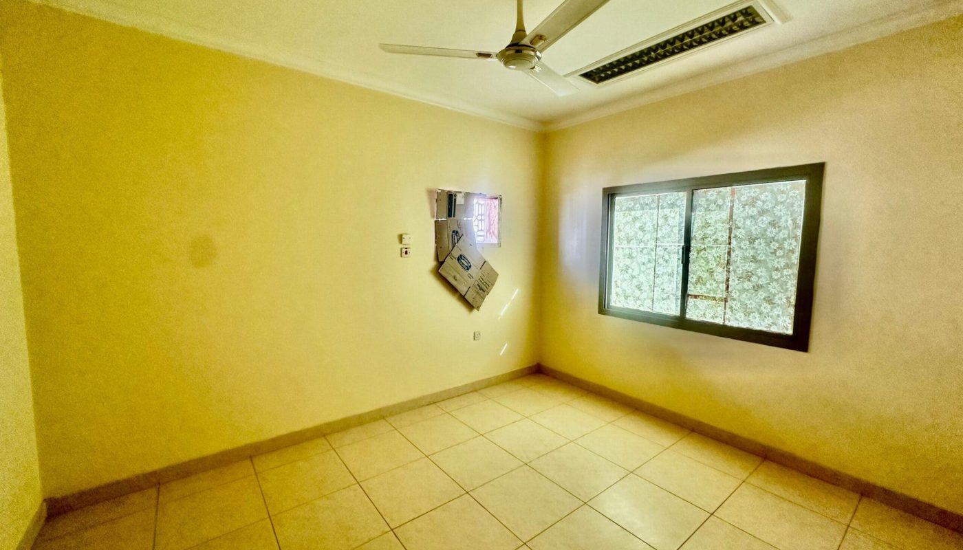 An empty room with tiled floors and a ceiling fan, located in the Janbyah area. The window features patterned glass, and a small air conditioning unit is installed on the wall. Ideal for those seeking two-bedroom apartments for rent.
