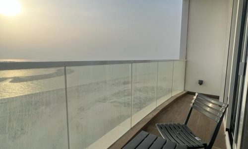 A balcony with a glass railing overlooking Bahrain Water Bay at sunset, furnished with a simple chair and a side table.