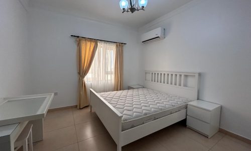 A minimalist bedroom in a flat for rent in Saar with a white double bed, desk, air conditioning unit, and a window with sheer curtains.