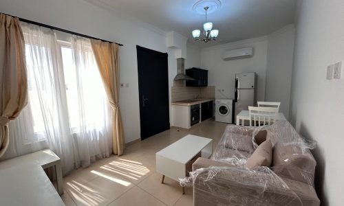 Modern living room interior in a flat for rent in Saar with furniture still wrapped in protective plastic, adjacent to a kitchen with appliances visible.