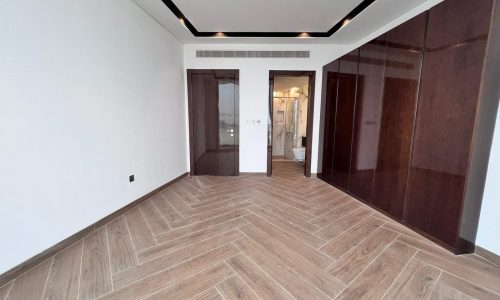 An empty apartment in Bahrain with a herringbone-patterned wooden floor and white walls, leading to a bathroom with an open door, overlooking the serene Water Bay Sea.