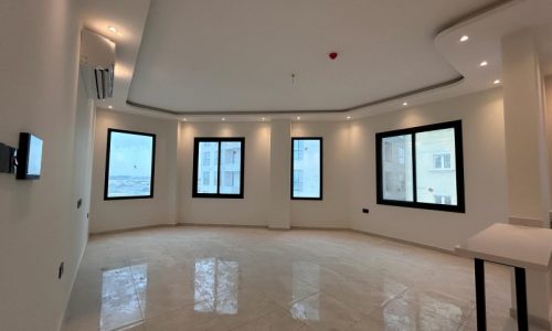 An empty apartment with polished floor tiles, white walls, multiple windows with black frames, recessed ceiling lights, and an unfurnished kitchen counter at the right is available for rent in the Janabiy