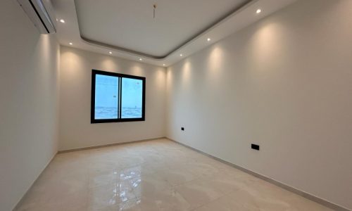 An empty apartment for rent in the Janabiyah Area with glossy tiled floors, white walls, recessed lighting, and a window with a view of the sky.