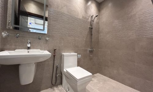 A modern bathroom interior in a Janabiyah area apartment for rent, with beige tiles, featuring a toilet, sink, mirror, and a wall-mounted towel warmer.