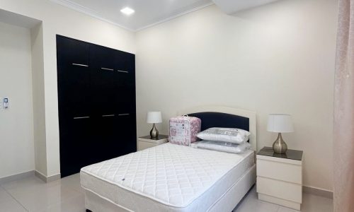 A neatly arranged bedroom featuring an unmade bed with pillows and a quilt on it, matching bedside tables with lamps, and a large black wardrobe against a white auto draft wall.