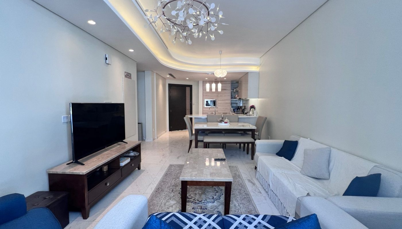 Modern living room with neutral-toned sofas, a coffee table, a flat-screen TV, and an open-plan dining area with a kitchen in the background. Contemporary lighting fixtures hang from the ceiling.