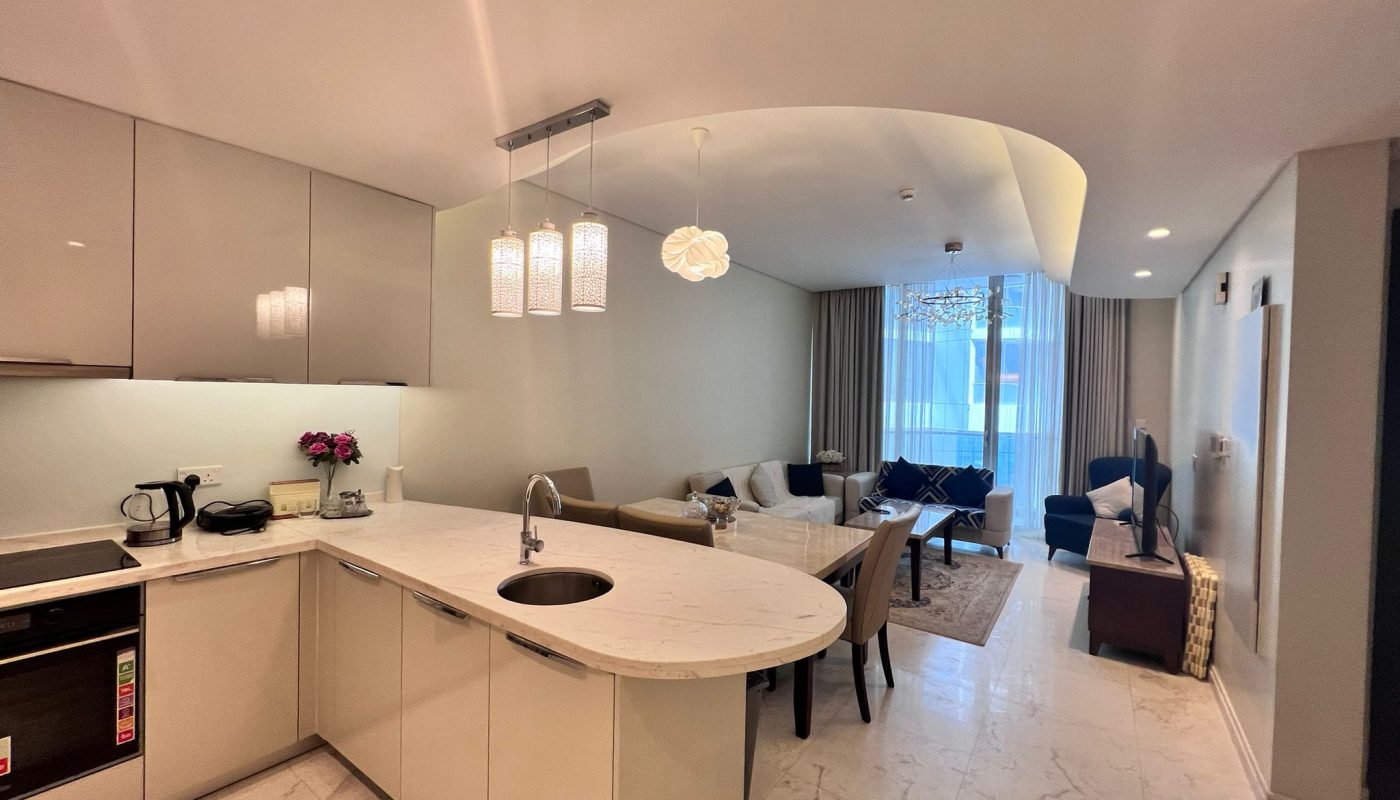 Modern open-plan kitchen and living area with marble floors, beige cabinets, pendant lighting, and large windows with sheer curtains.