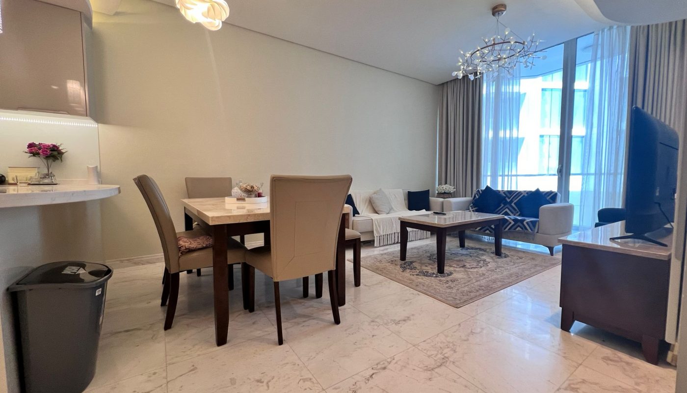 A modern open-plan living and dining area with a marble floor, beige chairs, a chandelier, large windows with curtains, and a TV on the right.
