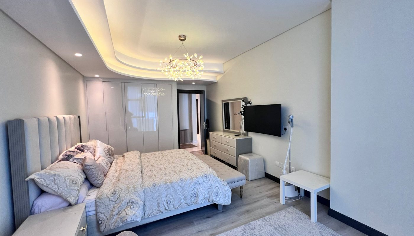 Modern bedroom with a double bed, decorative lighting, TV mounted on the wall, dresser, wardrobe, and neutral-colored decor.