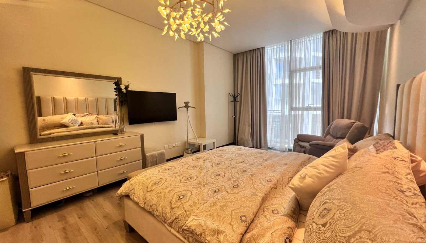 A modern bedroom with a large bed, dresser with mirror, wall-mounted TV, armchair by the window, and a decorative ceiling light fixture.