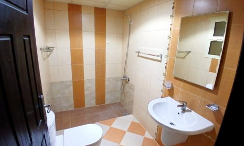 A clean, well-lit bathroom with beige and orange tiles, featuring a toilet, sink, shower, mirror, and an auto draft system.