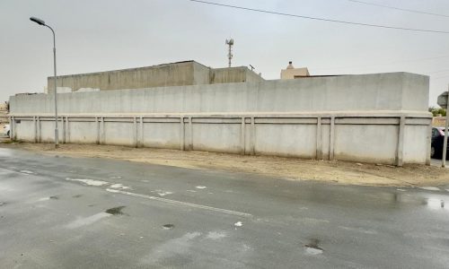 Overcast day featuring a wet street and a long concrete wall with the Aali single-story villa behind it, situated on a corner location.