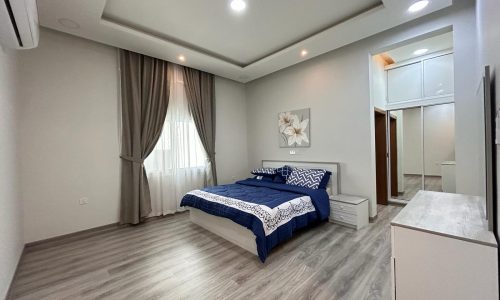 A modern, fully-furnished bedroom with a neatly made bed, hardwood flooring, and a wardrobe.