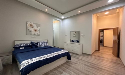 A modern bedroom in a fully-furnished apartment with a neatly made bed, white dresser, and wall art in a clean, well-lit space.