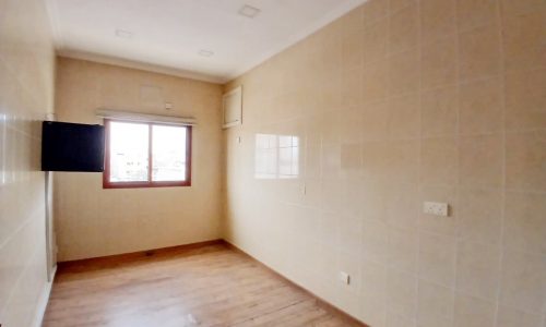 An empty room with beige tiled walls, wooden flooring, and a window letting in natural light through an auto draft.