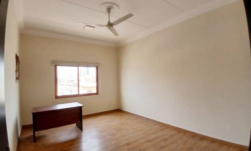 Empty commercial flat with wooden flooring, a ceiling fan, and a single brown desk near the window in Guful's prime location.