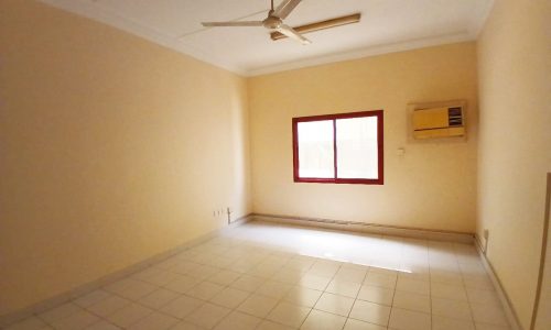 A sparsely furnished commercial flat in a prime location with tiled flooring, featuring a ceiling fan, an air conditioning unit, and a single window with a red frame.