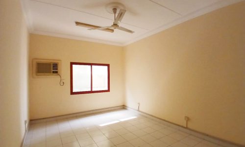 Empty room in a Commercial Flat in Guful, equipped with a ceiling fan, air conditioning unit, and a single window with a red frame, located in Prime Location.