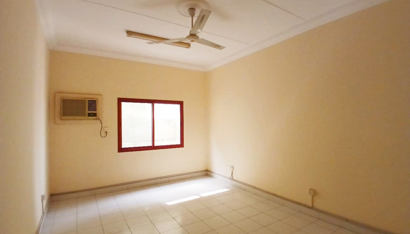 Empty room in a Commercial Flat in Guful, equipped with a ceiling fan, air conditioning unit, and a single window with a red frame, located in Prime Location.