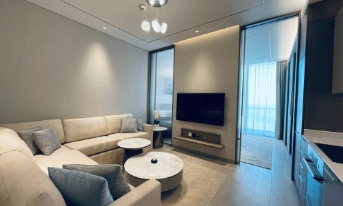 Modern living room in an amazing apartment for rent in Seef, featuring a beige sectional sofa, round coffee table, flat-screen TV, and sleek cabinetry, with ample natural light filtering through sheer curtains