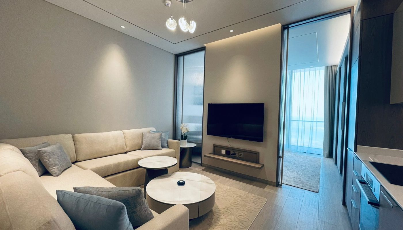 Modern living room in an amazing apartment for rent in Seef, featuring a beige sectional sofa, round coffee table, flat-screen TV, and sleek cabinetry, with ample natural light filtering through sheer curtains