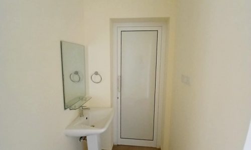 A minimalist bathroom in a brand new apartment in Hidd with a white sink, mirror, and a closed door.