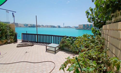 A sunny seaside terrace at a 3BR Villa in Amwaj with plants and a clear view of a calm blue sea and buildings across the water.