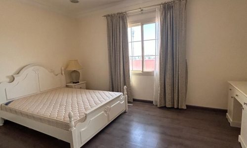 A fully-furnished bedroom within a 3 BDR villa in the Saar area, featuring a white bed frame, mattress, bedside lamp, and matching dresser. It has dark wood flooring and light