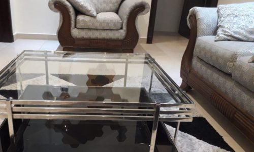 A living room featuring a glass coffee table in the foreground with a classic armchair and a sofa in the background, all placed on a patterned rug, available for rent in Busaiteen.