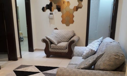 A living room in a Busaiteen flat for rent, featuring a patterned sofa and armchair, geometric wall art, a patterned rug, and two doors.