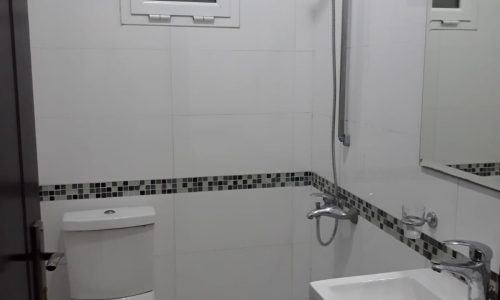 A small, clean bathroom in a flat for rent with a white toilet, sink, mirror, and a walk-in shower area with white tiles and black accents.