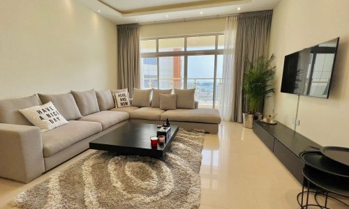 Modern living room featuring a large beige sectional sofa with decorative pillows, a dark coffee table, and a television, overlooking a city view through large windows.